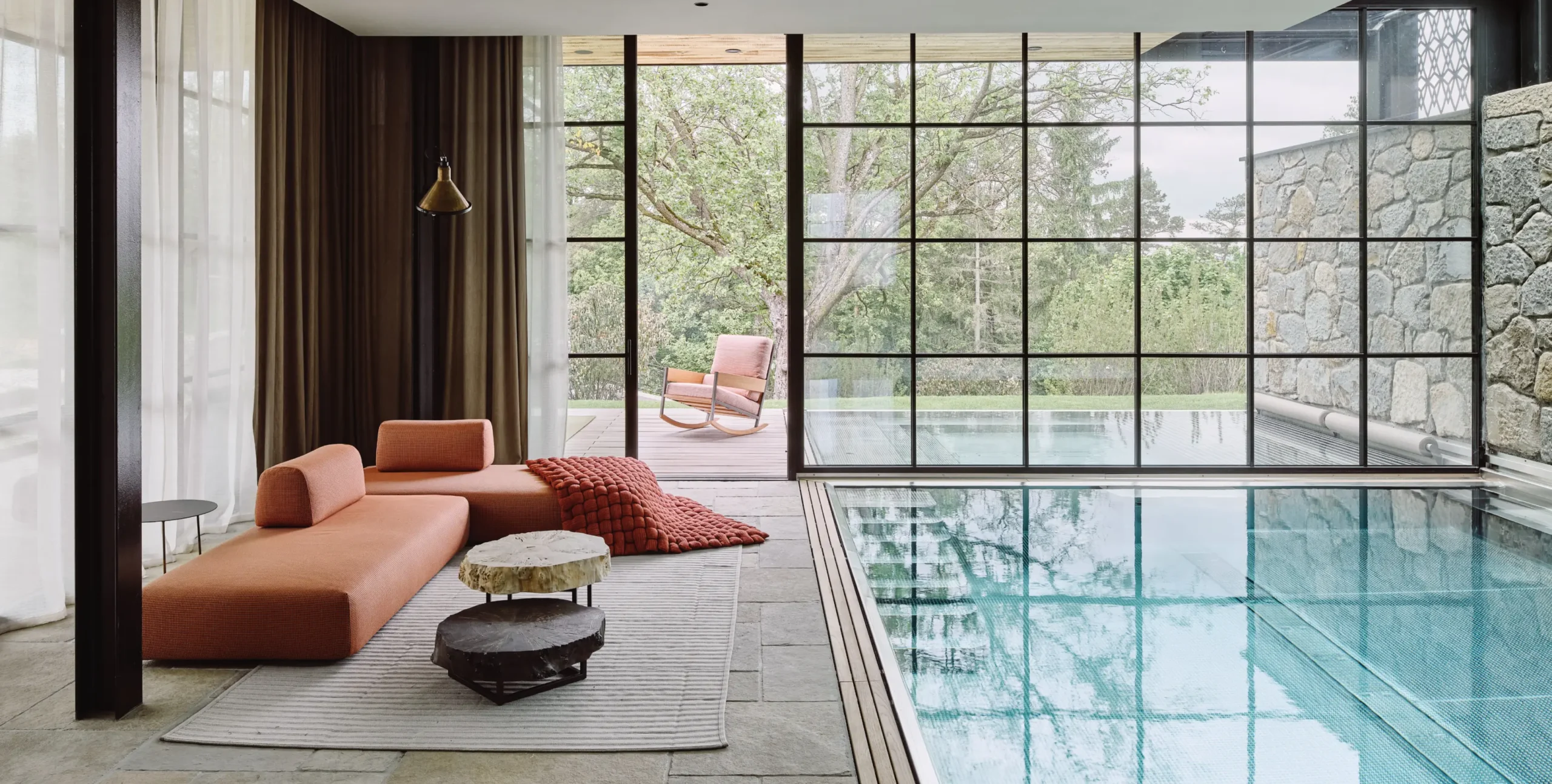 Luxuriöser Innenpoolbereich mit bodentiefen Fenstern, modernem Loungesofa in Erdtönen, dekorativen Kissen und Blick ins Grüne.