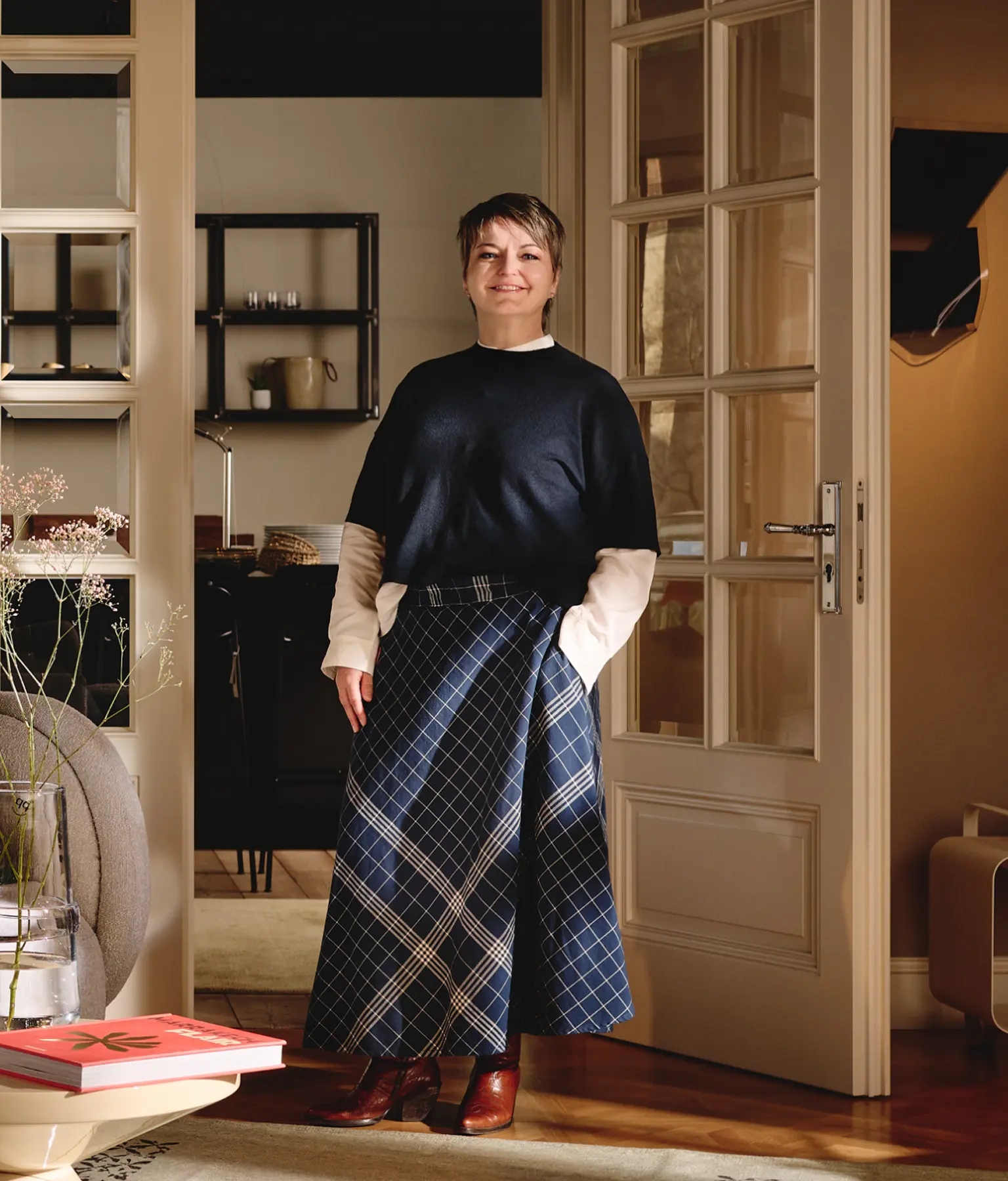 Stilvoll gekleidete Person mit kariertem Rock und dunklem Oberteil steht in einer elegant eingerichteten Wohnung mit offener Tür und warmem Licht