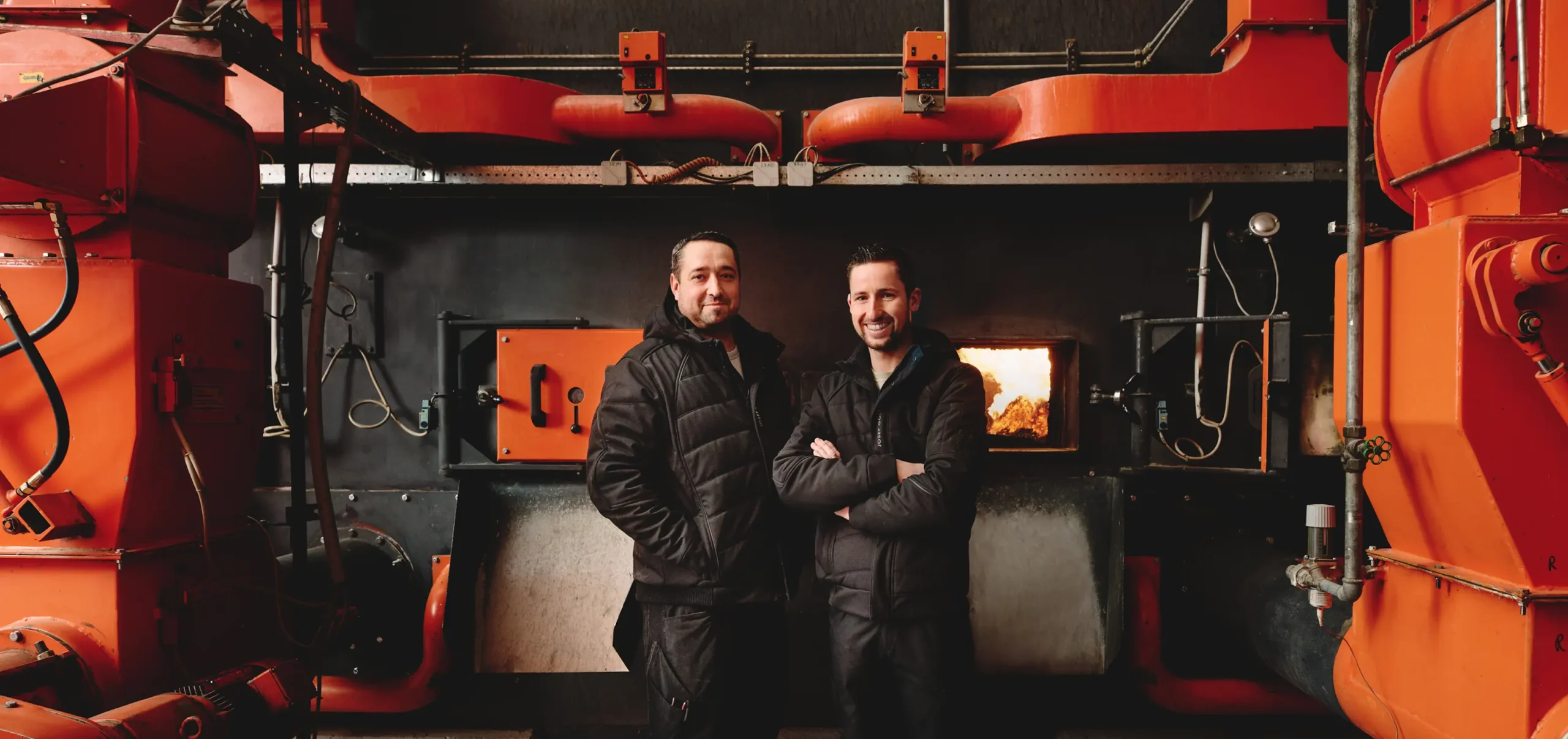 Zwei Männer in schwarzen Jacken stehen vor einer großen Industrieheizanlage mit orangefarbenen Rohren.
