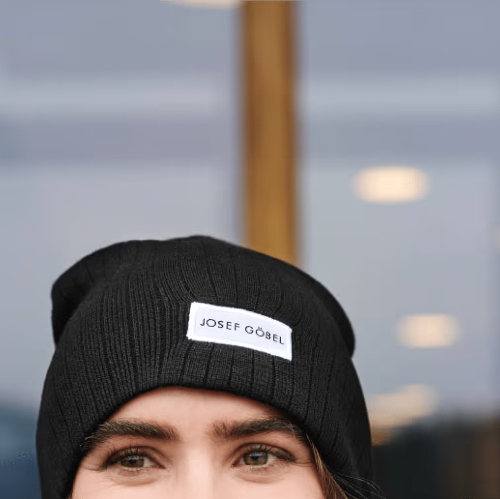Junge Frau mit schwarzer Strickmütze, auf der „Josef Göbel“ steht, vor einem verschwommenen Hintergrund mit warmem Licht.